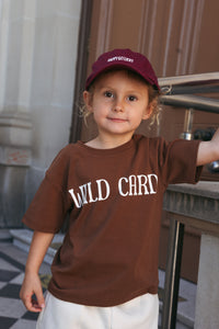 Child wearing a chocolate brown t-shirt with "WILD CARD" graphic, standing outdoors.