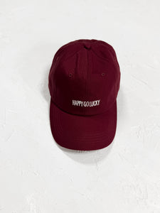 Burgundy cap with "Happy Go Lucky" embroidery, resting on a white surface.